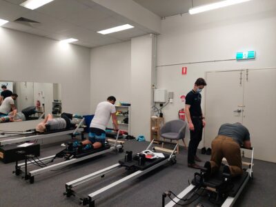 WhatsApp-Image-2021-09-08-at-11.40.18-AM-400×300-1 Group of people exercising on Pilates reformer machines under instructor supervision in a fitness studio.