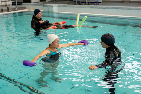 Three people participate in a water exercise session in an indoor swimming pool, using foam floats and hand weights.